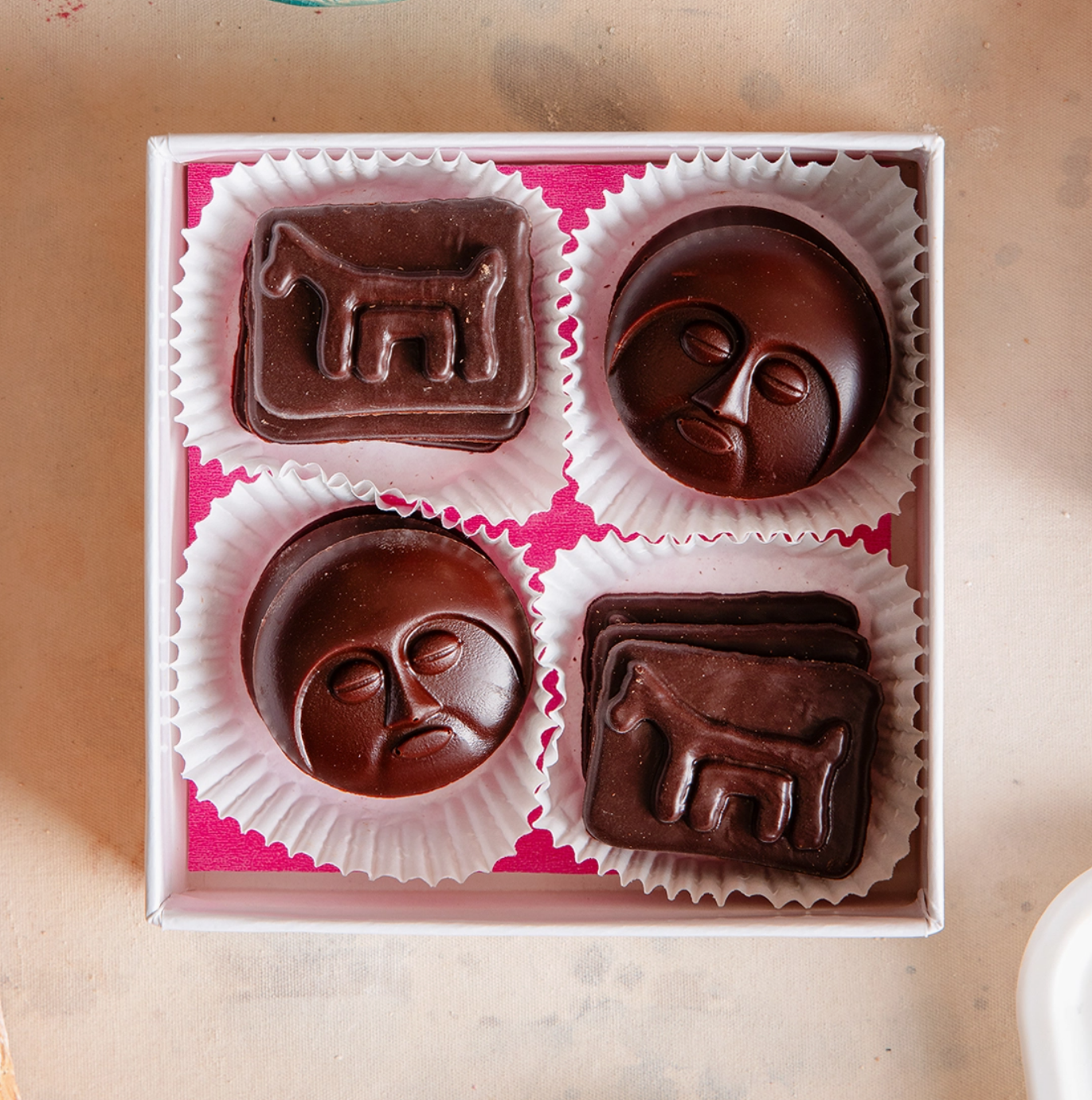 Celebrate! 4-piece vegan chocolate sampler featuring moon face and stylized creature sculptures, arranged in white paper cups inside a pink kraft bo