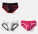 Three pairs of mens briefs arranged on a light gray background: black with red trim at the top left, white with black trim at the bottom left, and pink with white trim at the bottom right. Small red logo tabs are visible on each pair.