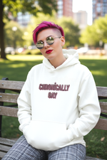 Person with bright pink hair wearing white Chronically Gay eco hoodie, sitting on a bench in urban park setting with plaid pants and reflective sunglasses