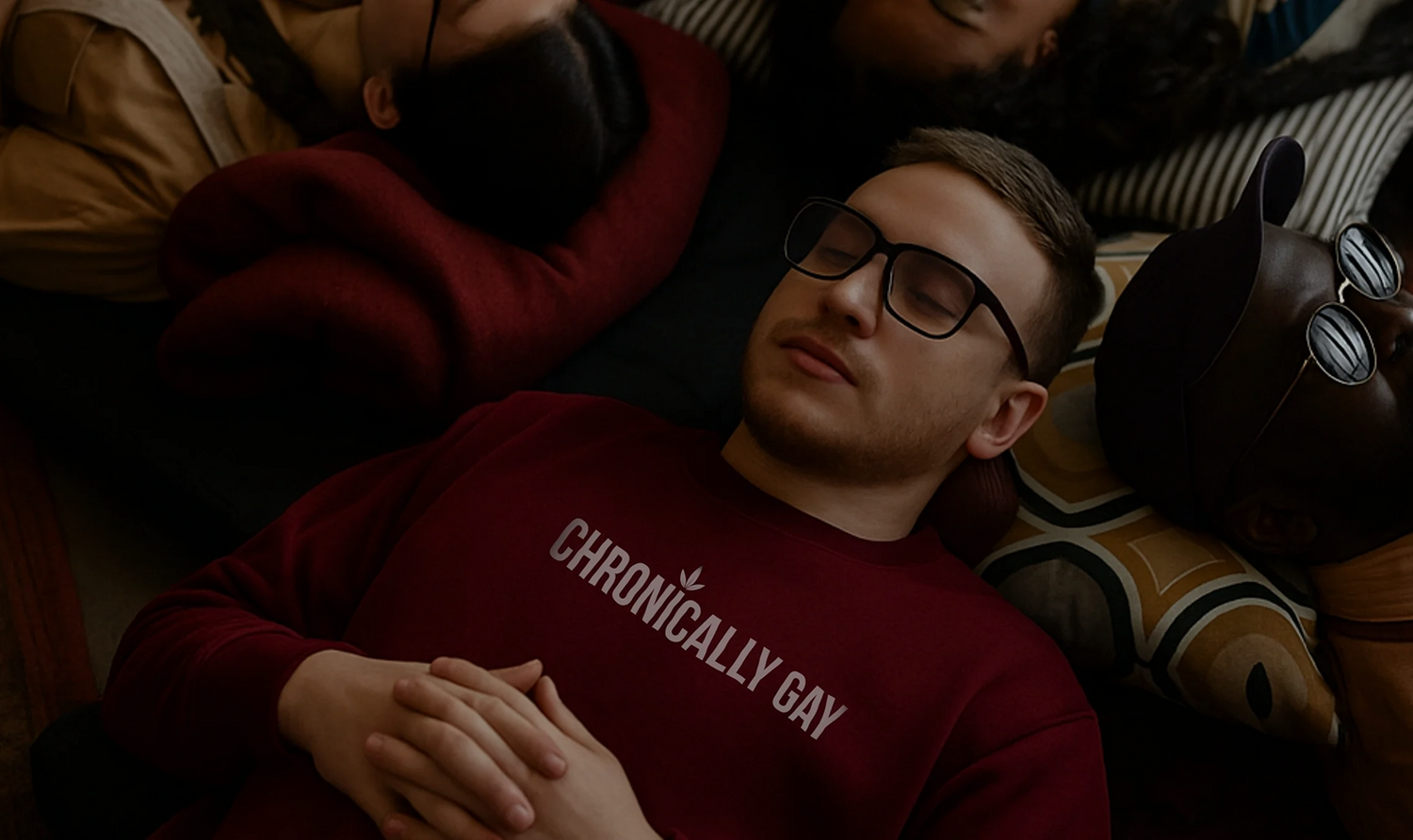 Group of people lying together on a couch, central figure wearing maroon “CHRONICALLY GAY” sweatshirt, part of queer fashion campaign