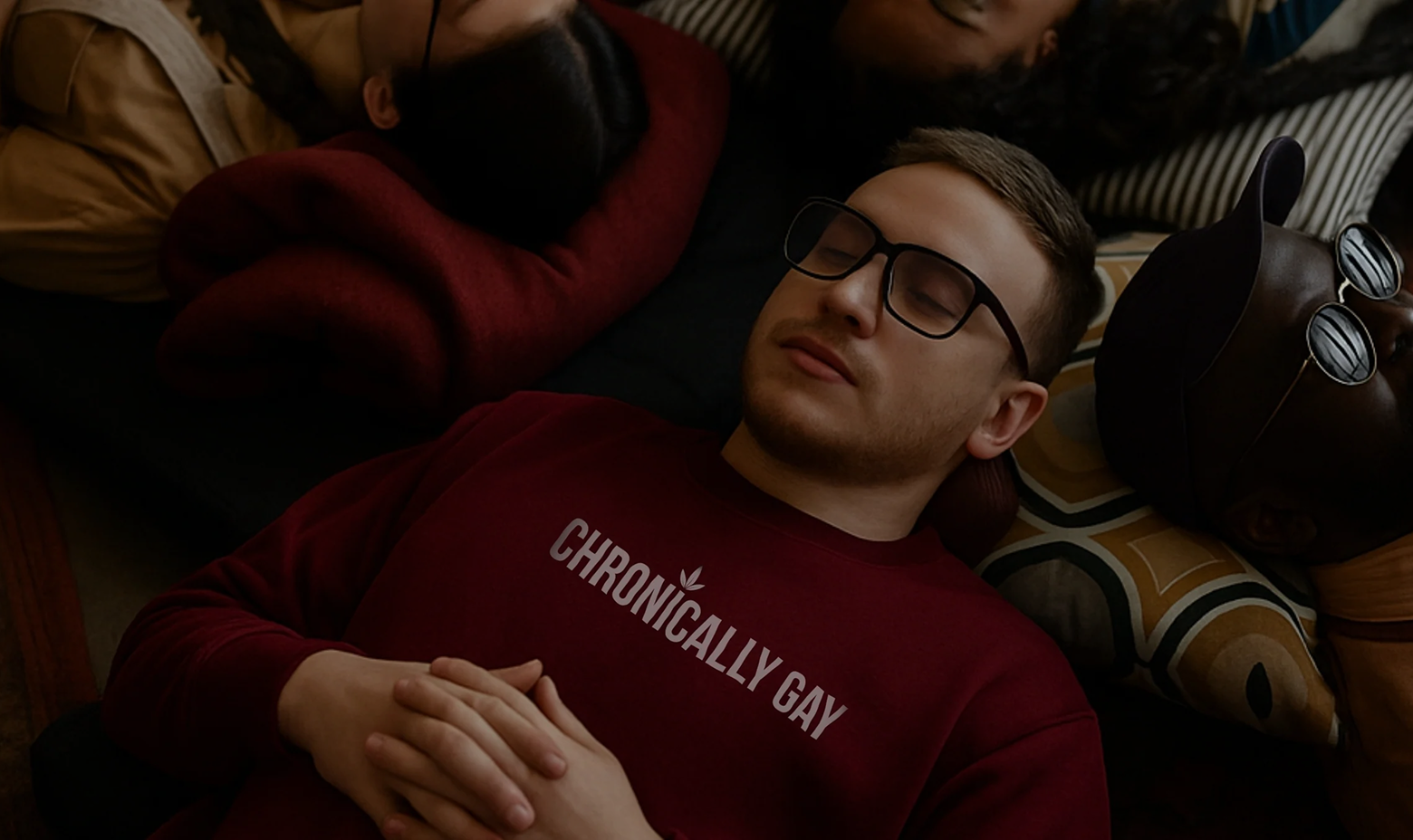 Group of people lying together on a couch, central figure wearing maroon “CHRONICALLY GAY” sweatshirt, part of queer fashion campaign